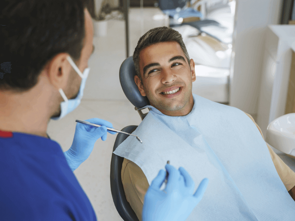 A cheerful young man smiling in a dental chair during a consultation with his male dentist, who is wearing blue gloves and a mask, in a modern dental clinic setting, portraying a positive and professional dental care experience.