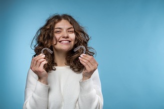 Person with curly hair in white sweater holding two clear dental aligners near face against light blue background.